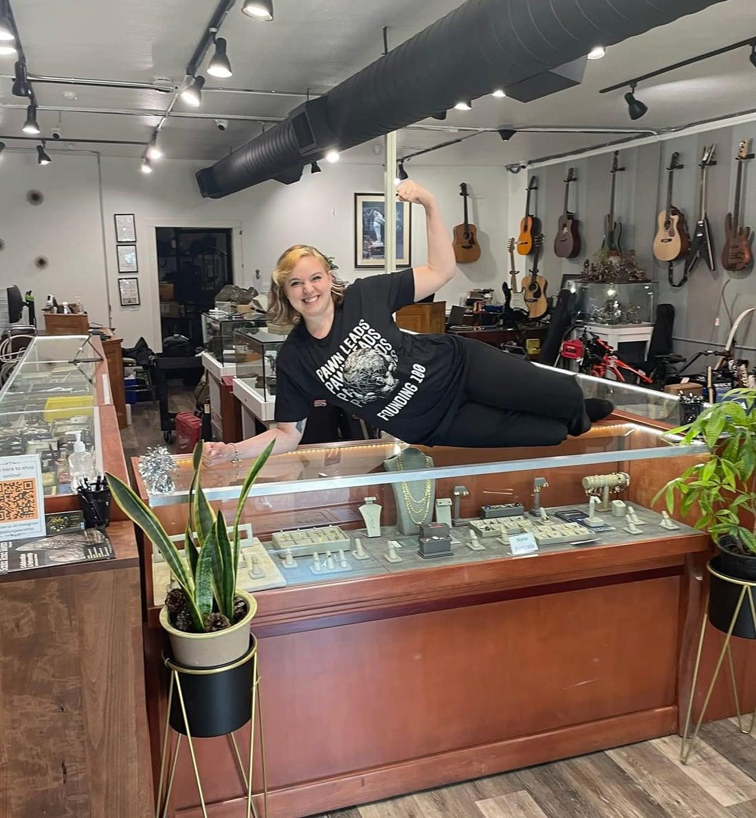 A smiling woman lying across a jewelry display case in a pawn shop, flexing, in a black Founding 100 shirt