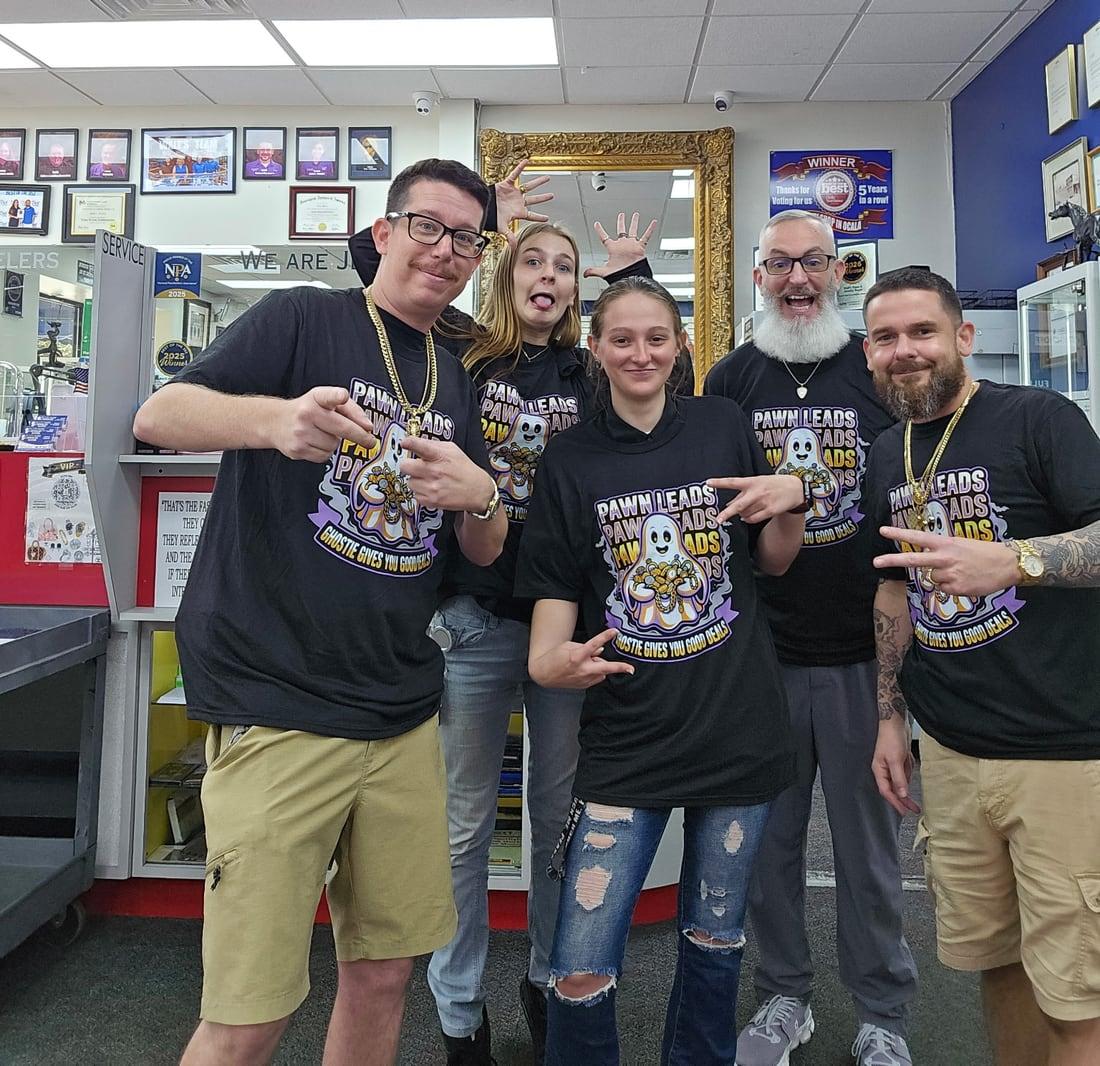 Five people at a pawn shop counter, all in the same black Ghostie Gives You Good Deals Pawn Leads shirt, pointing at the shirts and goofing for the camera
