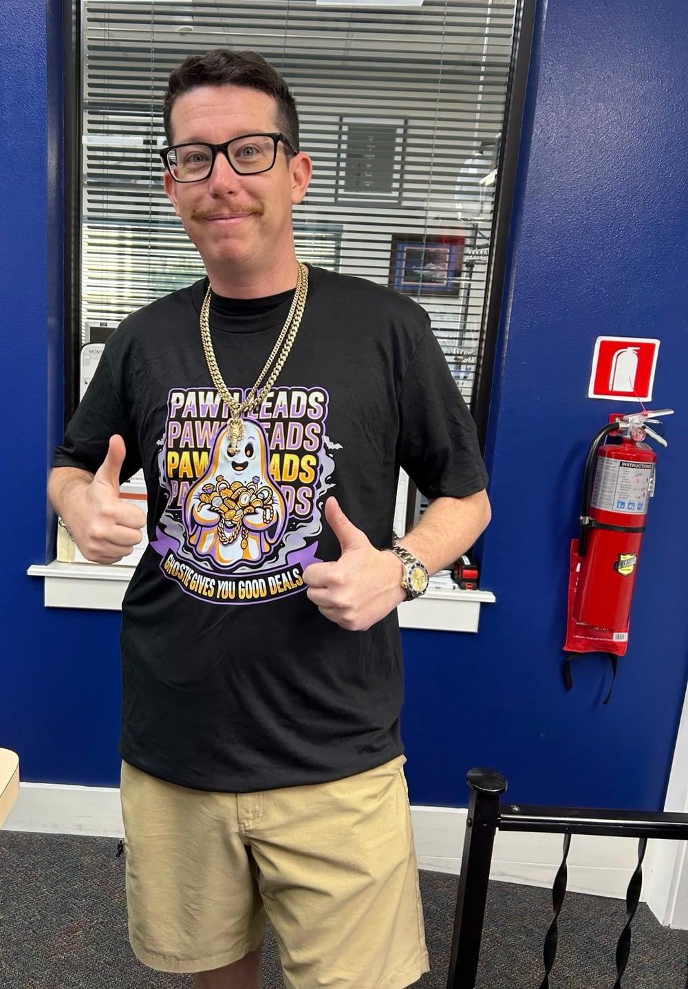 A man in a pawn shop giving two thumbs up in a black Pawn Leads shirt: a smiling ghost hugging a pile of gold and jewelry, with the line Ghostie Gives You Good Deals