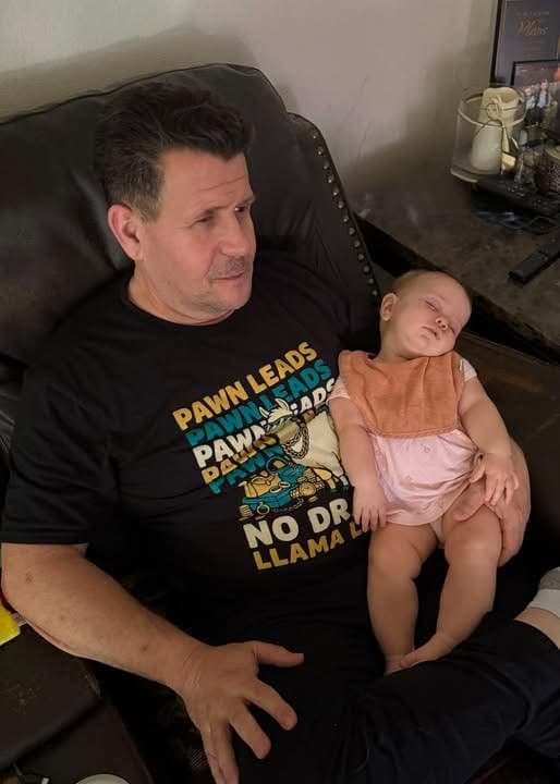 A man in a recliner with a sleeping baby on his lap, wearing a black No Drama Llama shirt