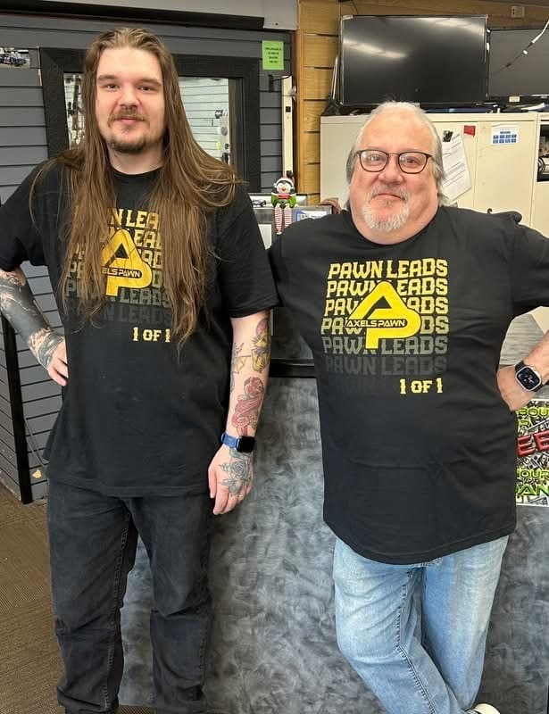 Two men behind a pawn shop counter in matching black 1 of 1 shirts with yellow Pawn Leads lettering