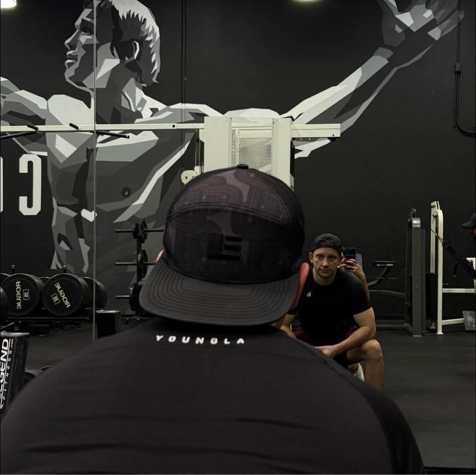 Jeff Trimble at the gym, seen from behind, facing a mirror under a bodybuilder mural